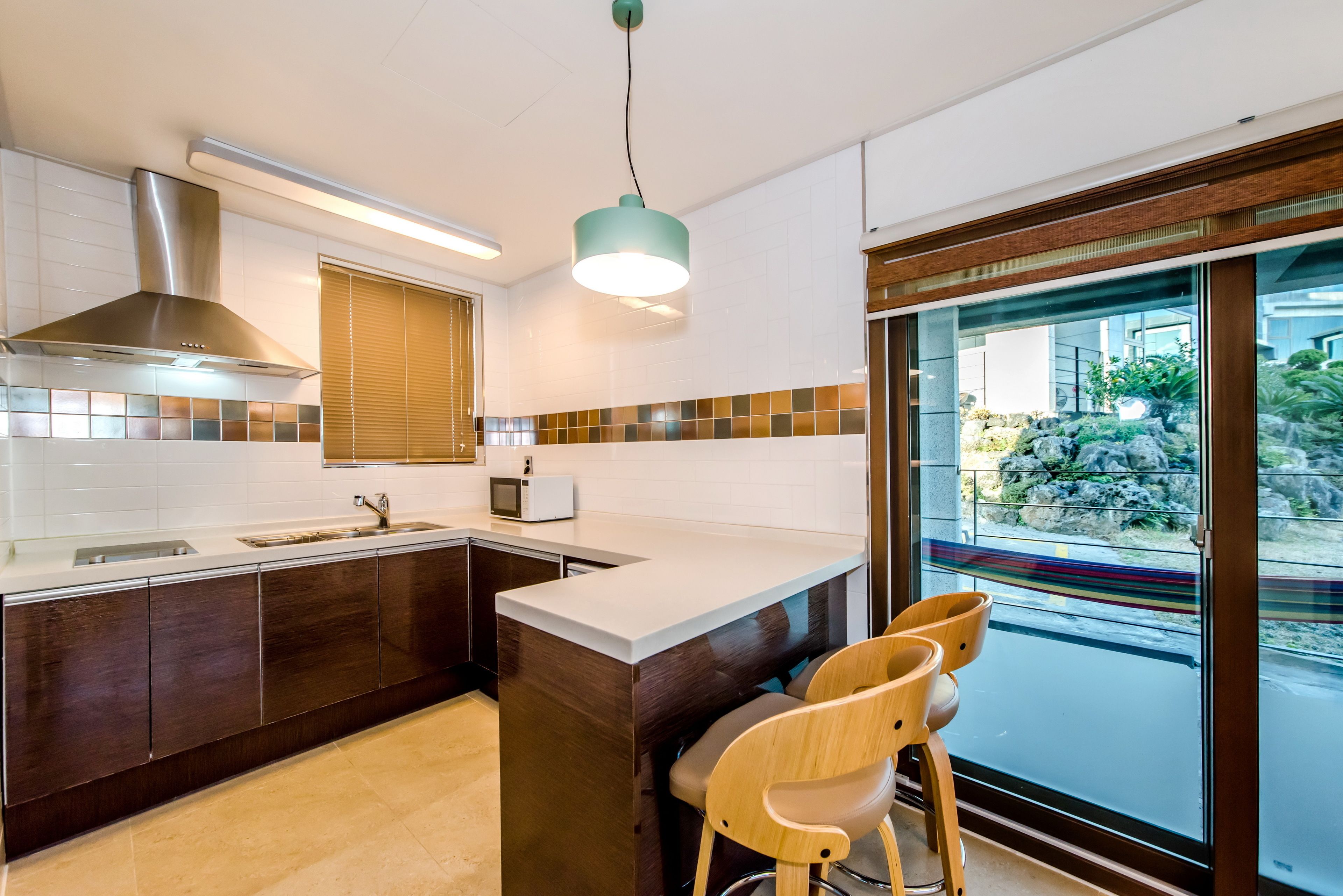 A modern kitchen with a large island, dark cabinetry, a stainless steel range hood, and a view through a glass door.