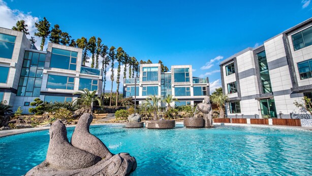 A modern hotel with a pool featuring a rock sculpture, surrounded by palm trees and modern architecture. - Donnaekohill Resort (Seogwipo)