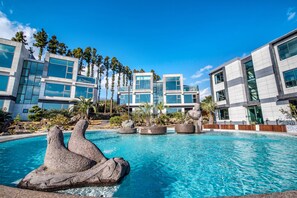 Un hotel moderno con piscina que presenta una escultura de roca, rodeado de palmeras y arquitectura contemporánea.