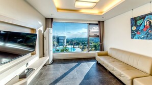 A modern living room with a large TV, a sofa, a painting, and a view of a swimming pool and buildings. - Donnaekohill Resort (Seogwipo)