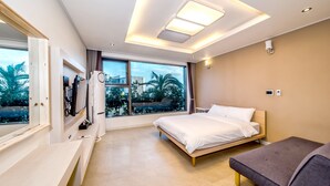 A modern hotel room with a large bed, a desk, and a view of palm trees through large windows. - Donnaekohill Resort (Seogwipo)