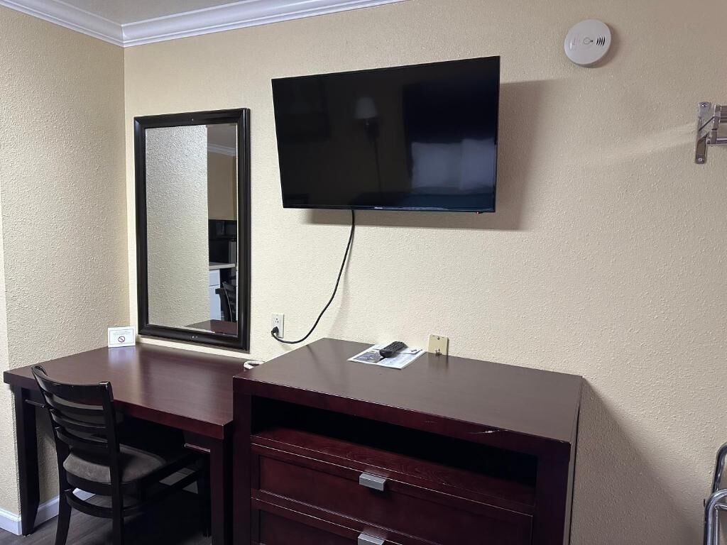 Habitación de hotel con televisor de pantalla plana montado en la pared, un escritorio de madera con una silla y un espejo.