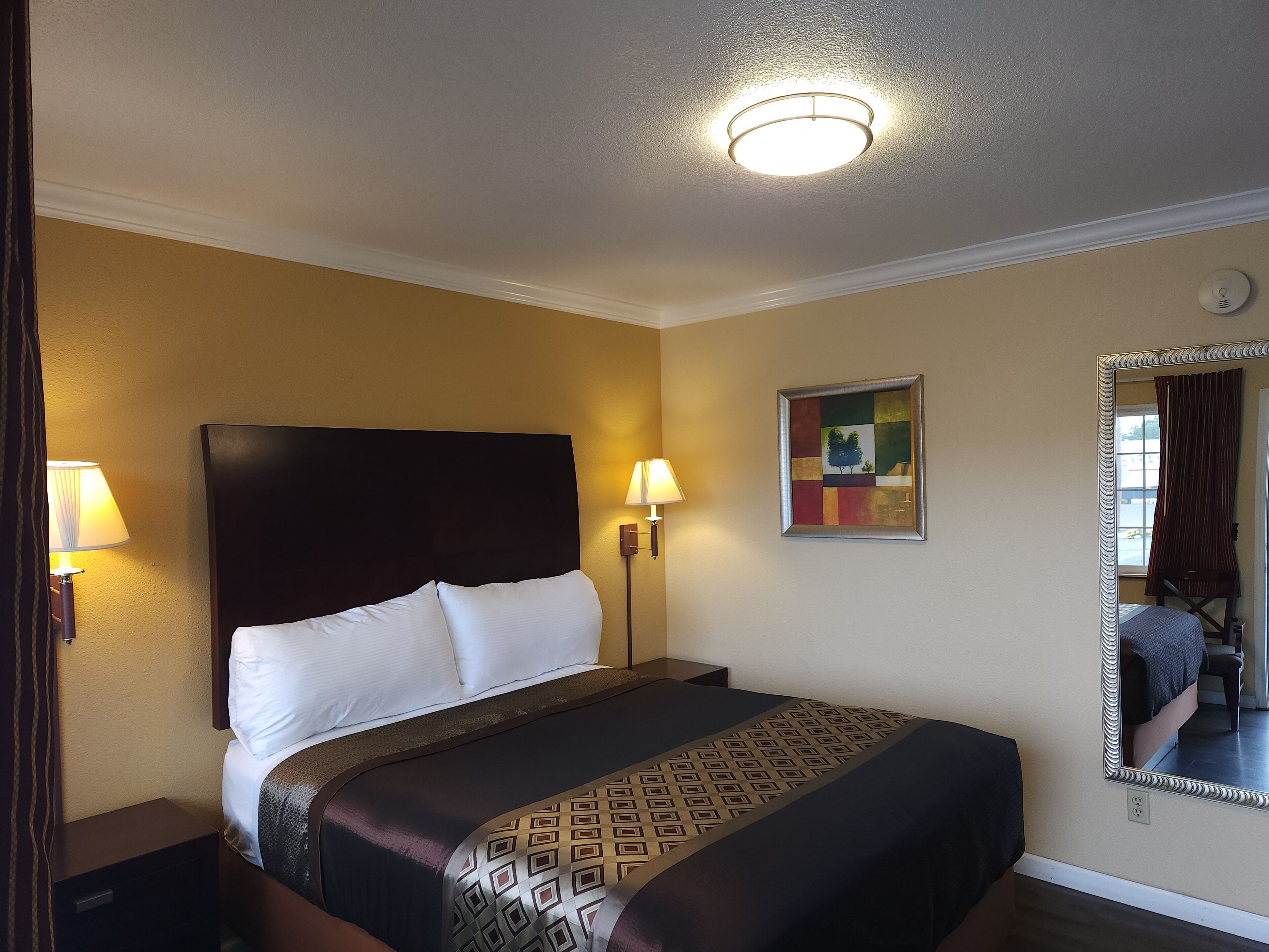 A hotel room with a large bed, two bedside lamps, a ceiling light, a mirror, and a painting on the wall.