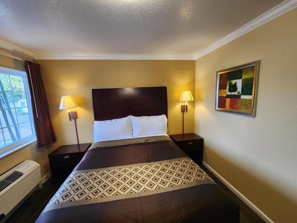 A hotel room with a large bed, two bedside lamps, a window with curtains, and a painting on the wall.