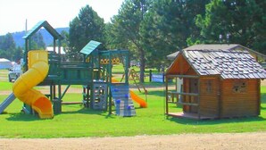 A playground with a slide and climbing structures, surrounded by trees and a small wooden cabin. - Rush No More RV Resort and Cabins - Campground (Sturgis)