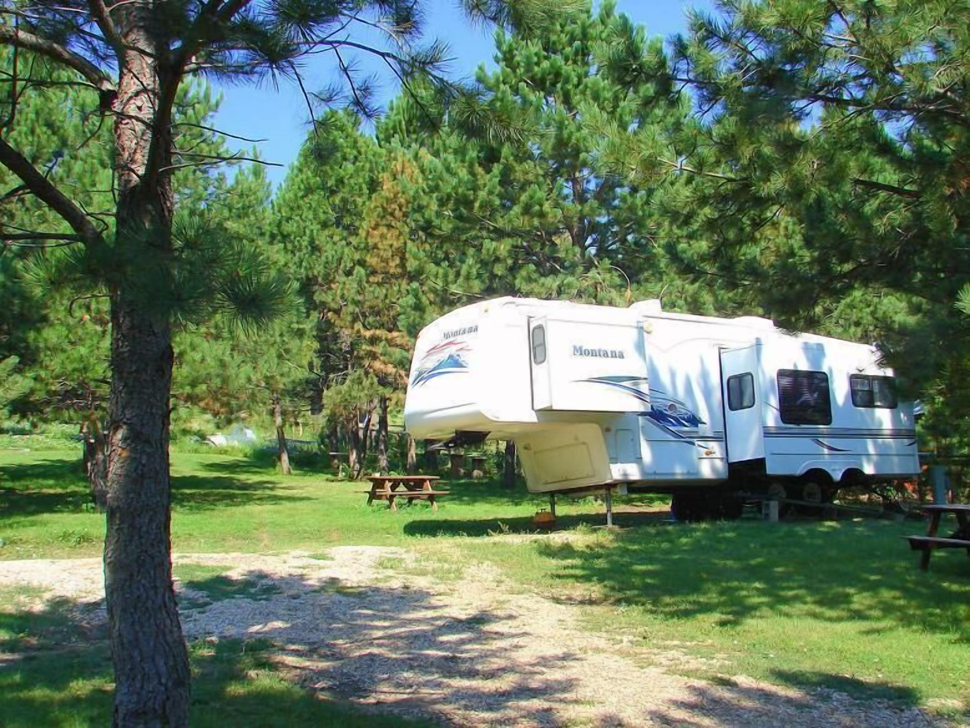 Una caravana blanca con el logotipo 'Montana' estacionada en un área boscosa con mesas de picnic.