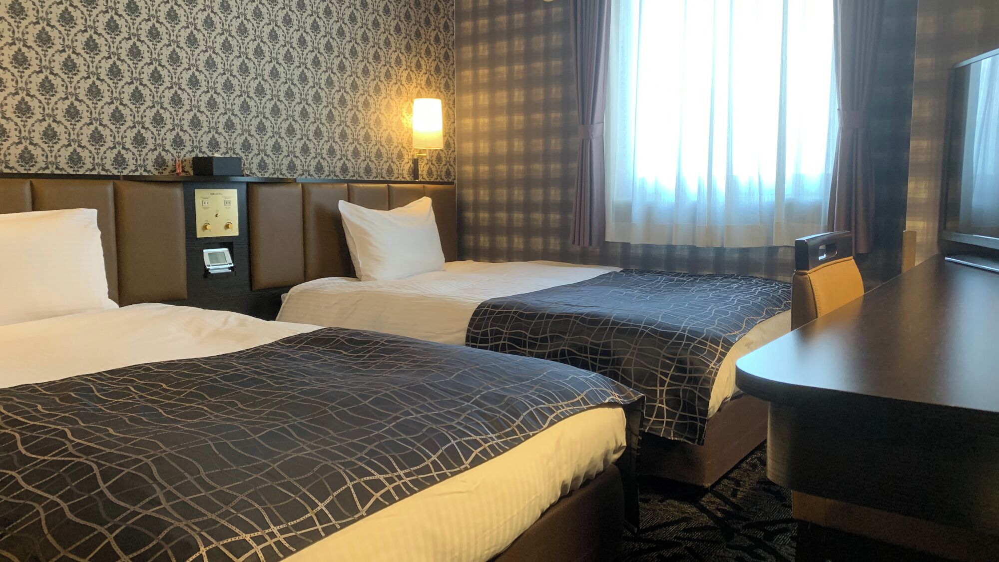 Hotellihuone, jossa on kaksi sänkyä, työpöytä, lamppu, kello ja ikkuna, jossa on verhot.