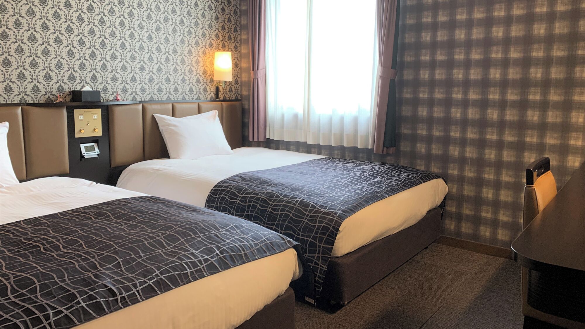 Hotellihuone, jossa on kaksi sänkyä, yöpöytä, lamppu ja ikkuna, jossa on verhot.