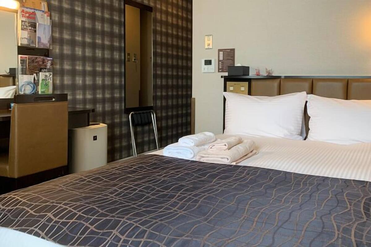 A hotel room with a bed, a desk, a chair, a wardrobe, and a wall-mounted lamp.