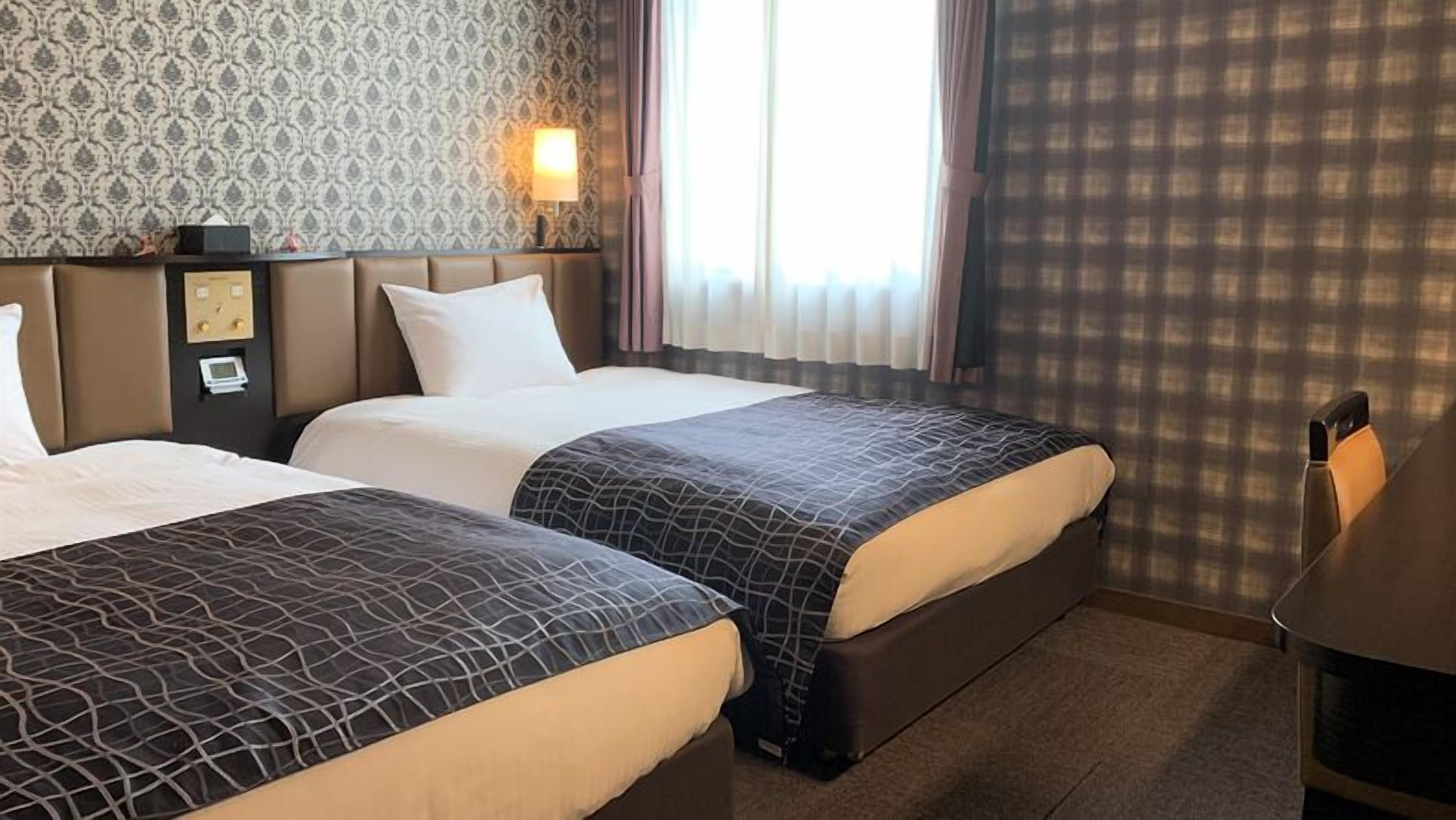Hotellihuone, jossa on kaksi sänkyä, sängynpääty, jossa on säätimet, yöpöytä ja ikkuna, jossa on verhot.