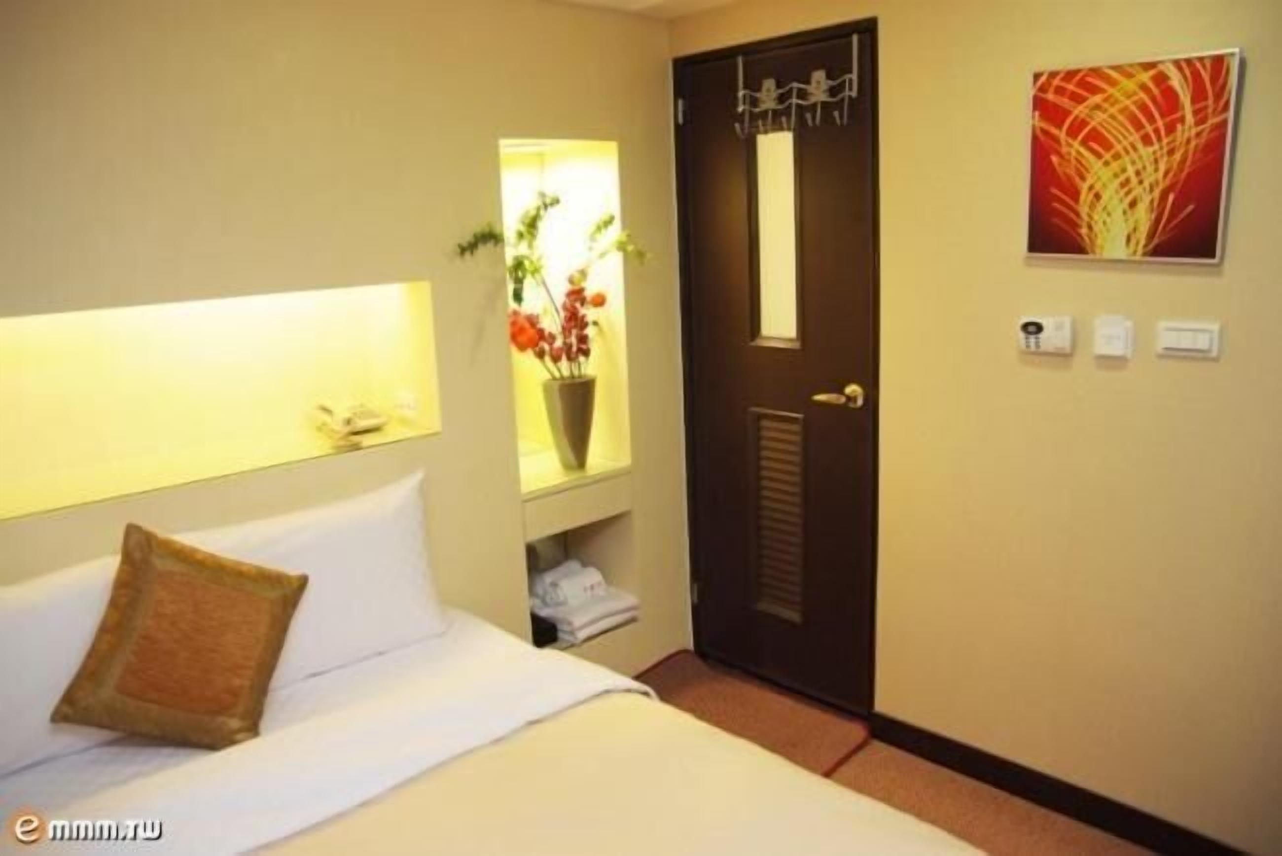 Una habitación de hotel con cama, mesita de noche con lámpara, puerta y estante con planta en maceta.