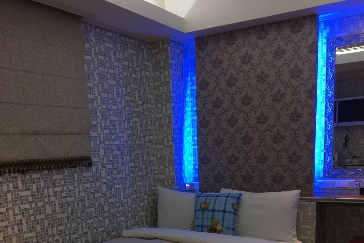 Un dormitorio con una cama grande, una ventana con iluminación azul, papel tapiz estampado y una mesita de noche con una lámpara.