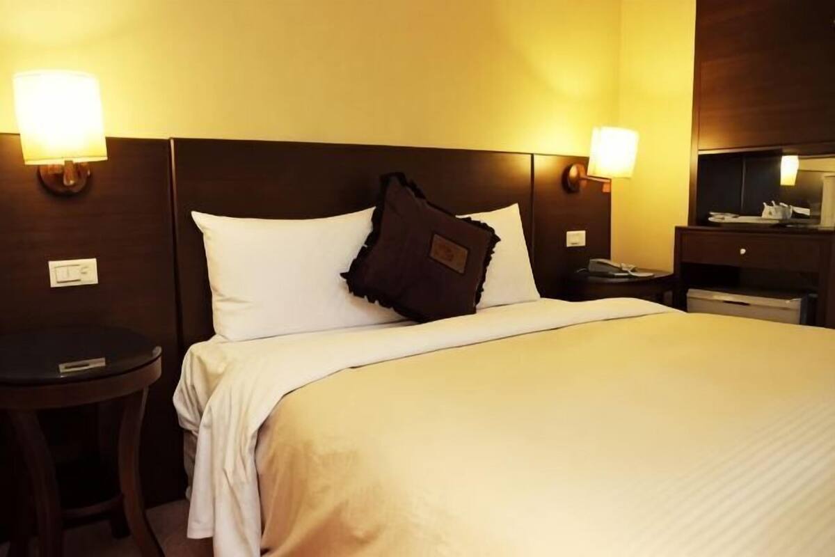 Una habitación de hotel con cama, mesitas de noche, lámparas y un pequeño mueble.