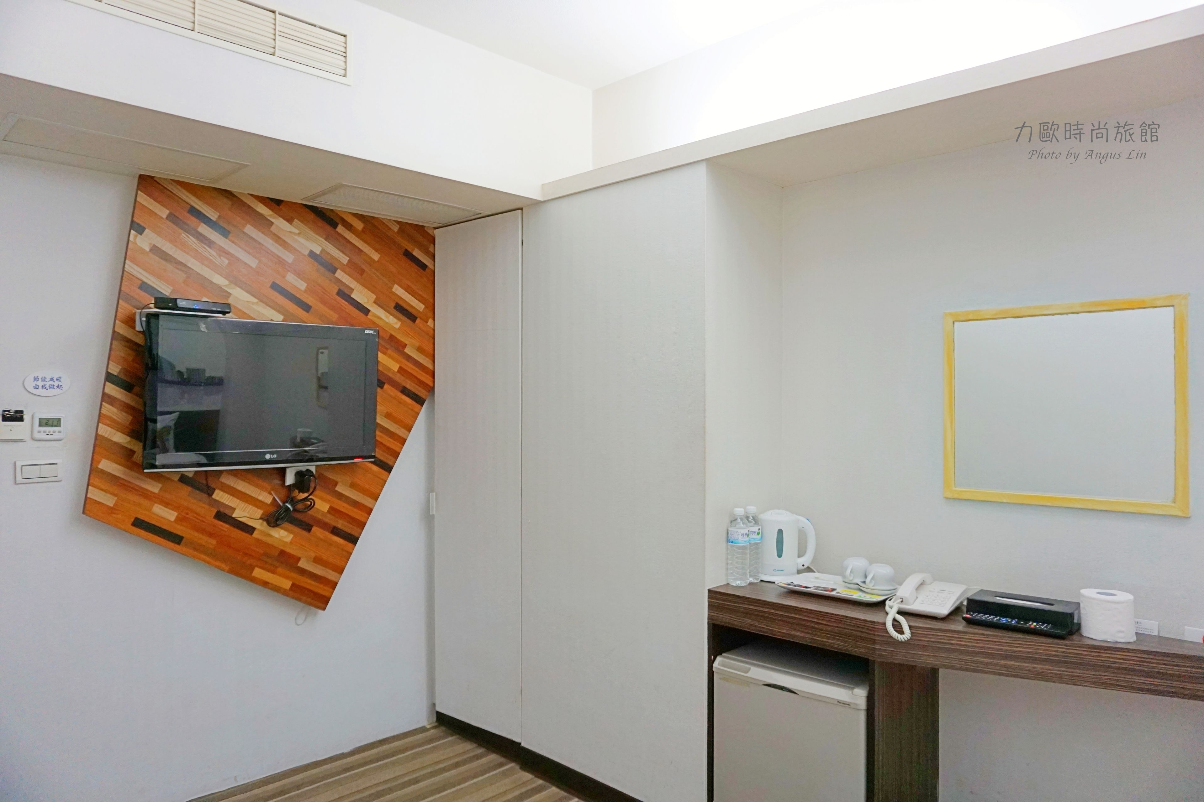 Habitación de hotel con televisor de pantalla plana montado en una pared de madera, un escritorio pequeño con teléfono, hervidor de agua y una botella de agua.