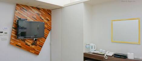 Habitación de hotel con televisor de pantalla plana montado en una pared de madera, un escritorio pequeño con teléfono, hervidor de agua y una botella de agua.