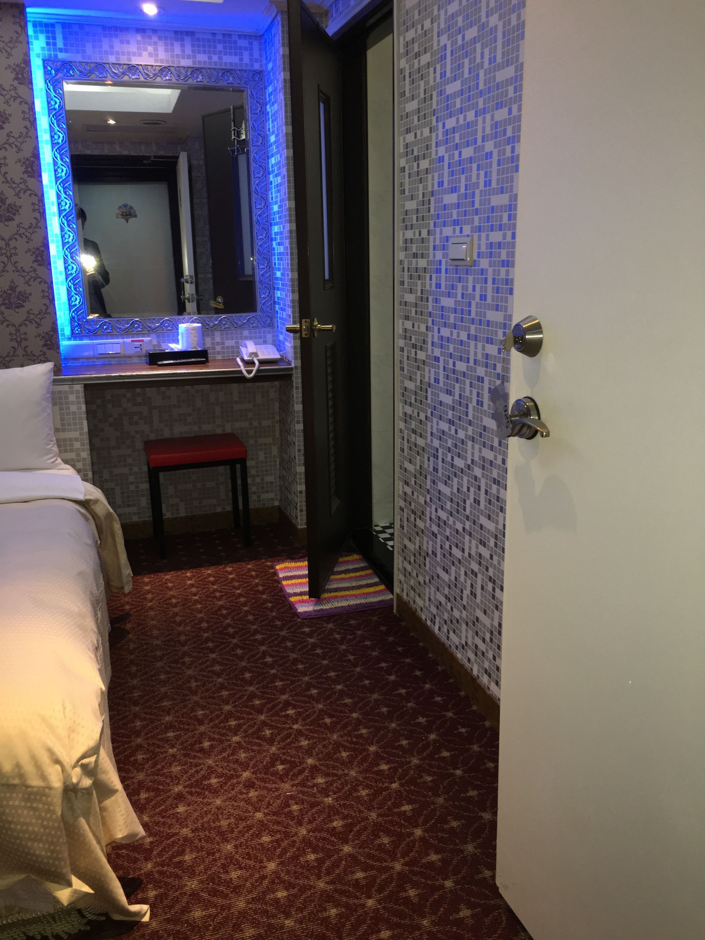 Quarto de hotel com cama, uma pequena mesa com telefone, espelho e porta.