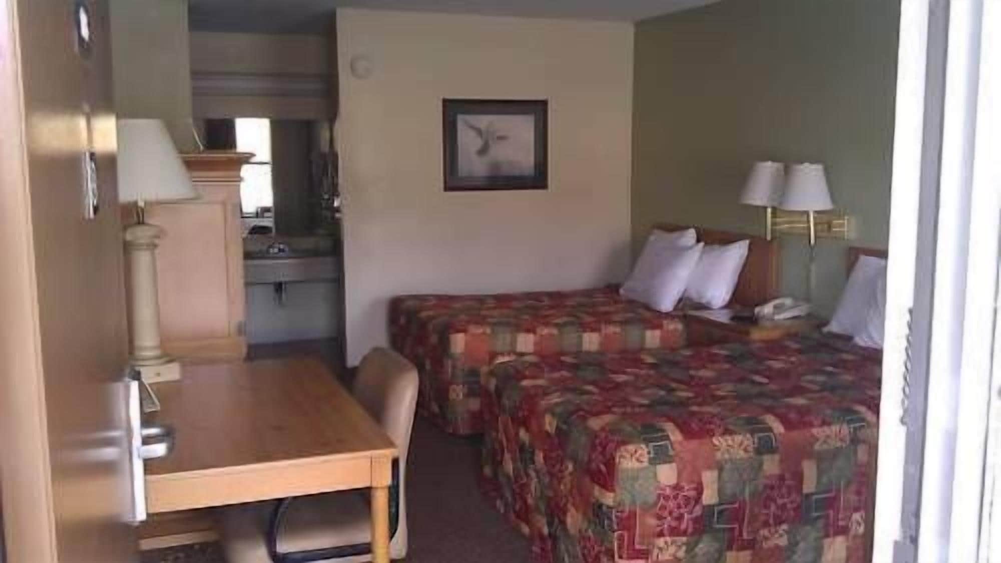 A hotel room with a bed, a desk, and a chair. There is a small kitchenette in the background.