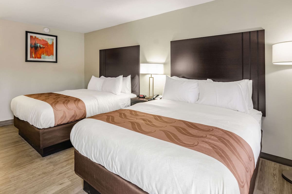 A hotel room with two beds, a nightstand with a lamp, a painting on the wall, and a wooden floor.
