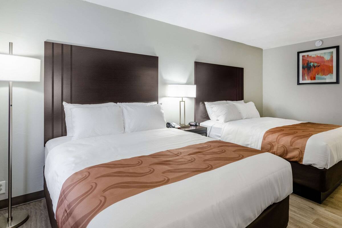 A hotel room with two beds, a bedside table with a lamp, and a framed abstract painting on the wall.