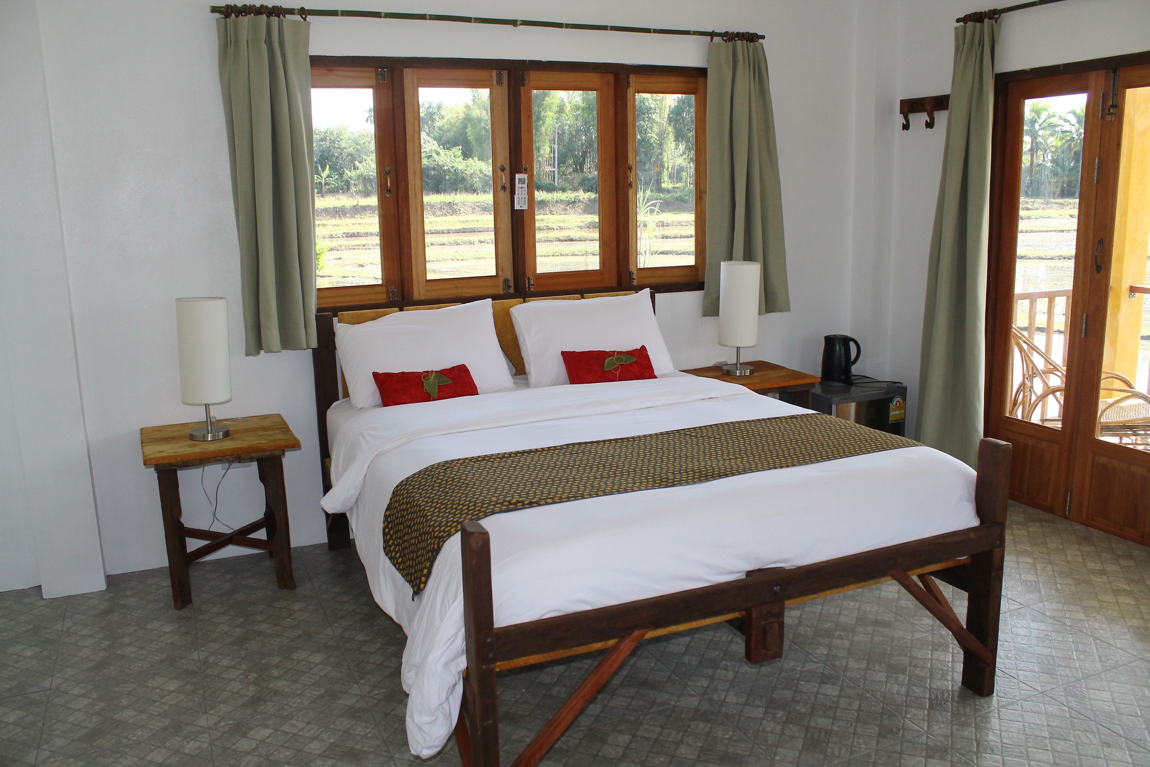 A large bed with white linens and red pillows, two bedside tables with lamps, and a view of a garden through large windows.