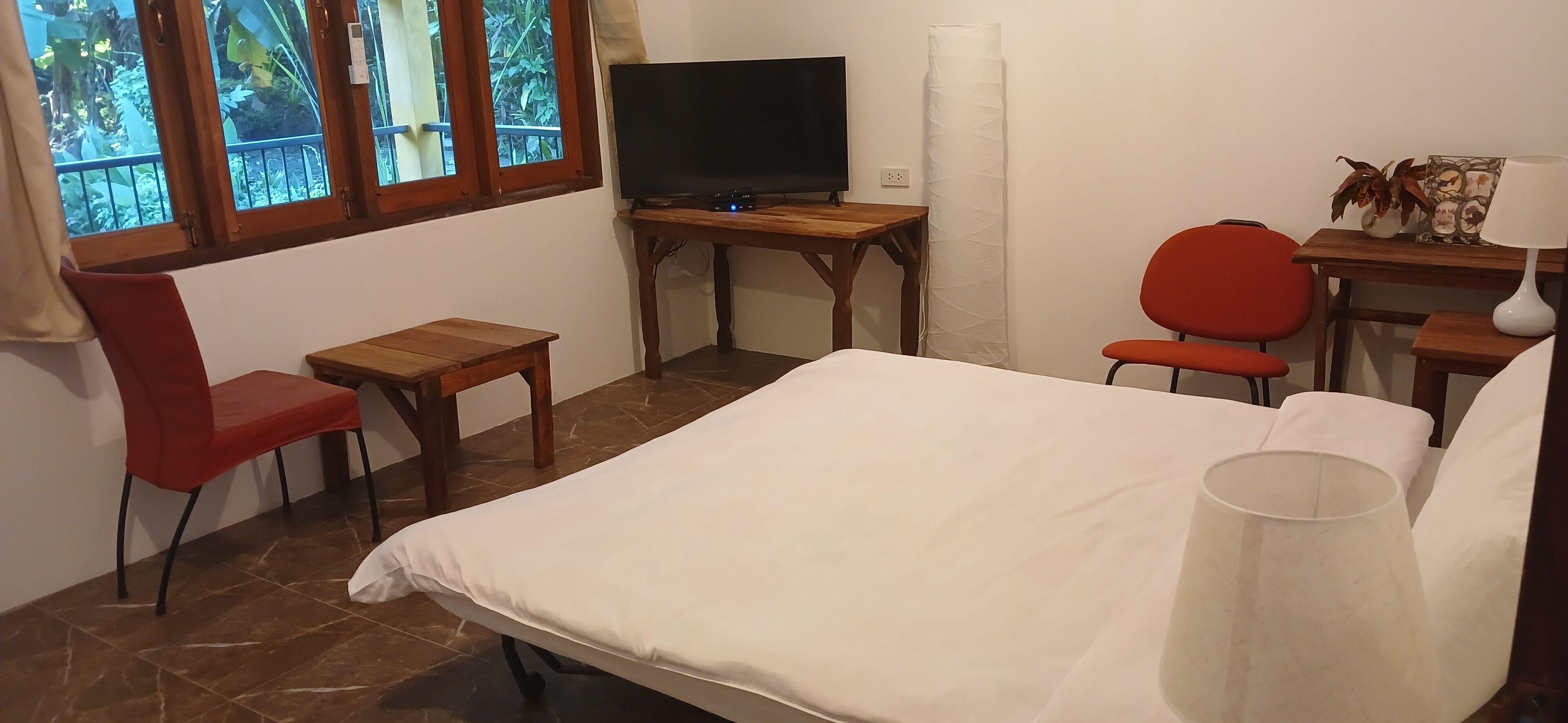 A hotel room with a bed, a desk with a TV, a red chair, and a window with a view of greenery.