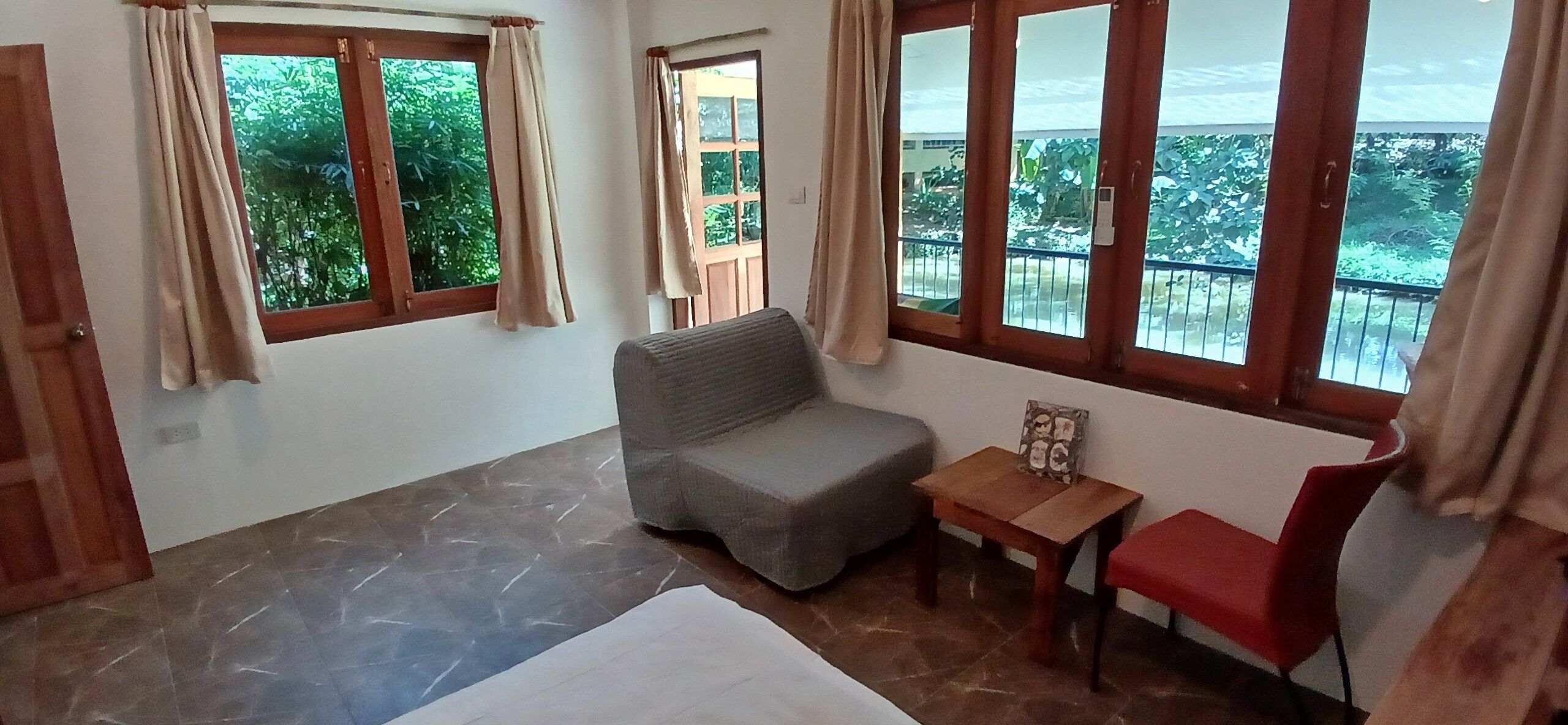 Quarto com cama, sofá cinza, mesa pequena com um travesseiro e uma cadeira vermelha. Há janelas com cortinas, e é visível uma varanda com guarda-corpo através das janelas.