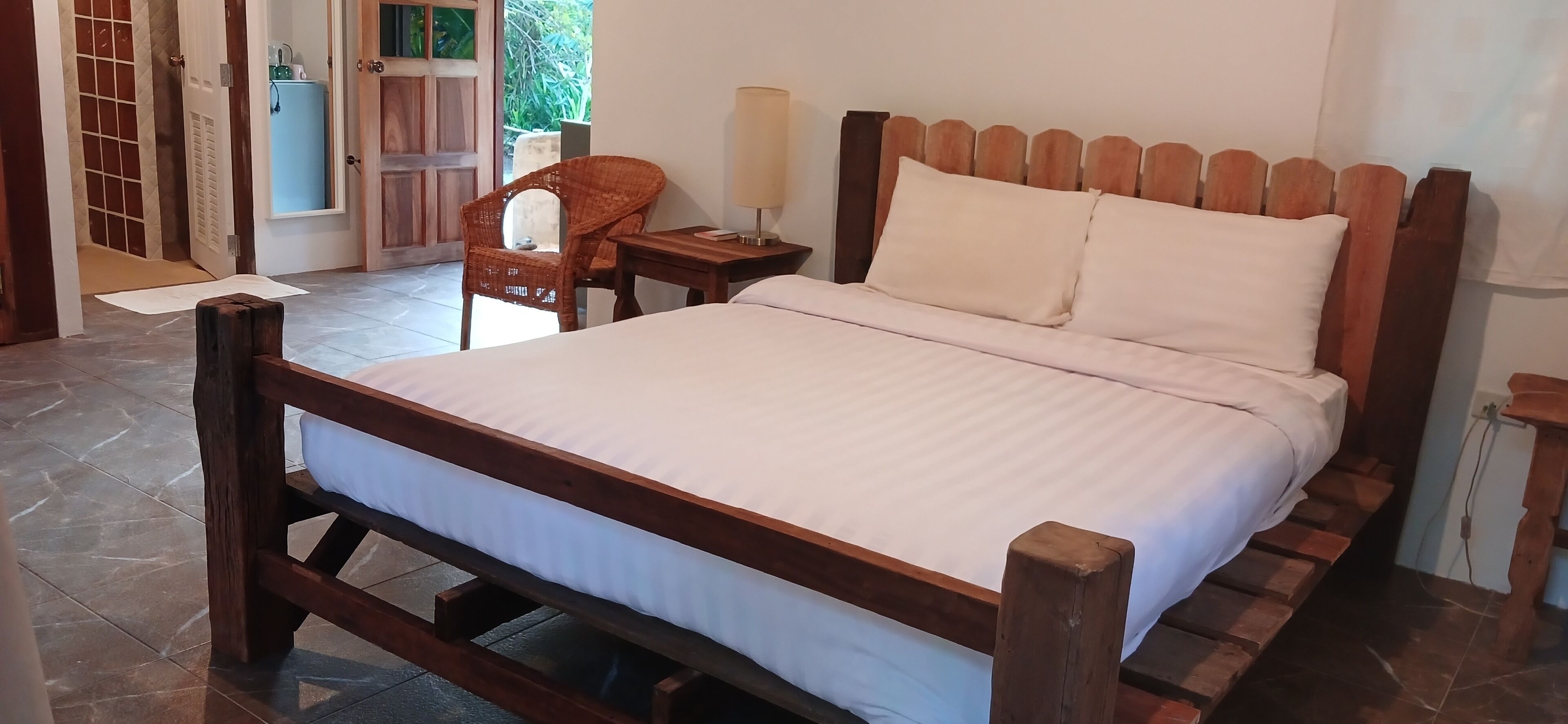 A wooden bed with white bedding, a wooden nightstand with a lamp, and a wicker chair in a room with tiled flooring and a door leading outside.