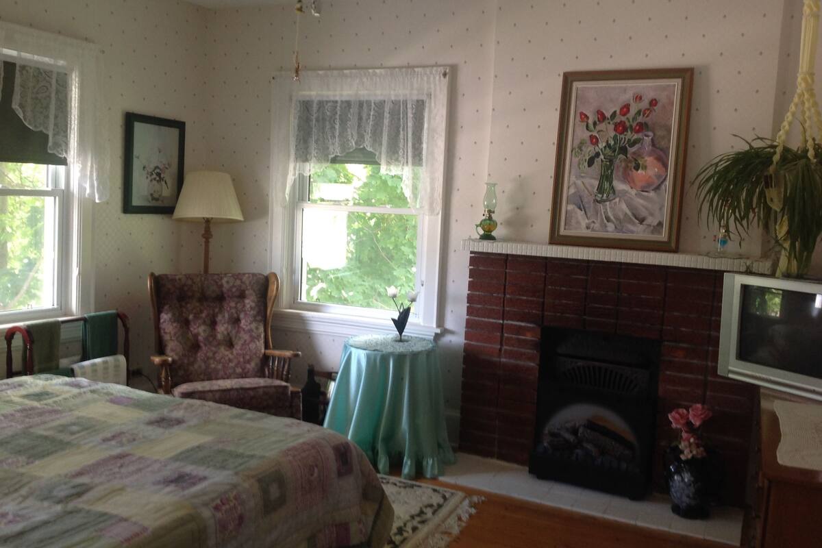 Un dormitorio acogedor con una cama, una silla, una chimenea y una ventana con cortinas de encaje.
