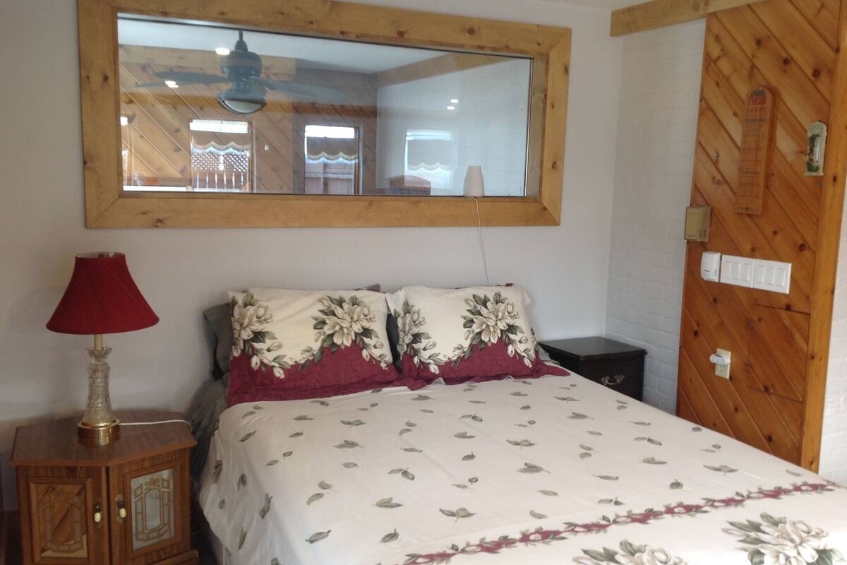 Un dormitorio con cabecera de madera, un espejo grande sobre la cama, una mesita de noche con lámpara y una colcha estampada con motivos florales.