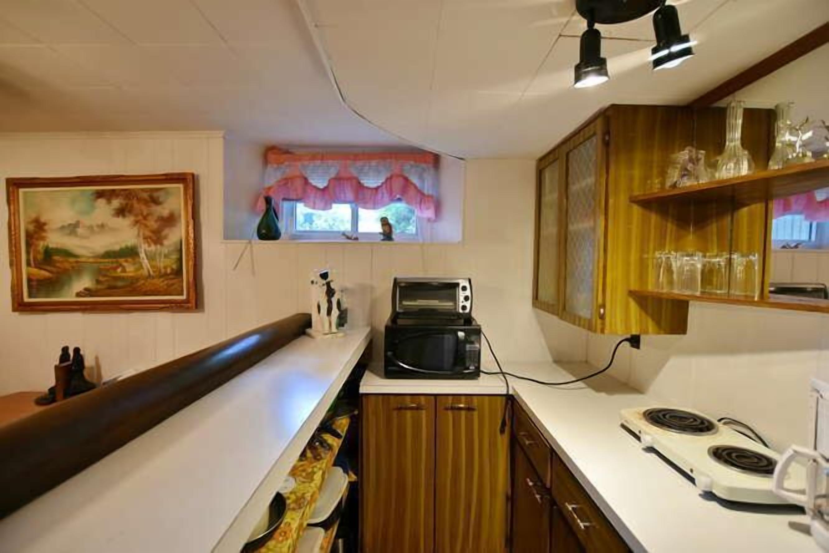 Une cuisine compacte équipée d’un four à micro-ondes, d’un grille-pain et d’une cuisinière. On y trouve une fenêtre avec un rideau rose, un tableau accroché au mur et des placards en bois.