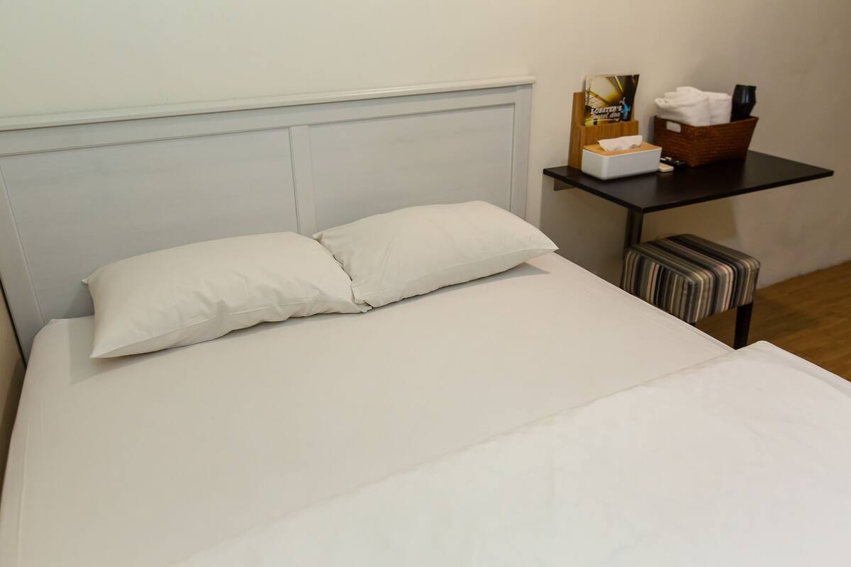 A neatly made bed with white linens and pillows, a bedside table with a basket of toiletries, and a striped bench.