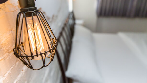 Un primo piano di un lampadario in metallo con una lampadina a filamento scoperto, fissato a una parete bianca.
