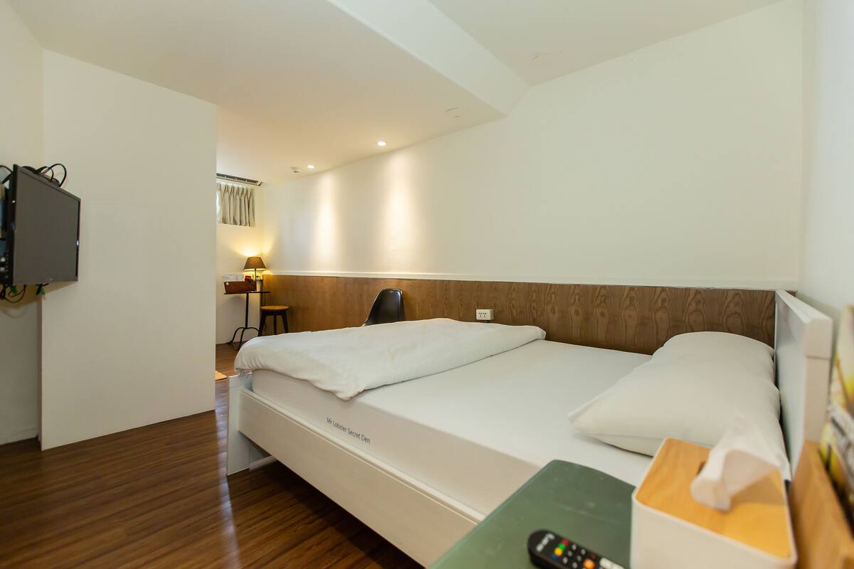 A hotel room with a bed, a bedside table with a remote control, a lamp, and a TV mounted on the wall.