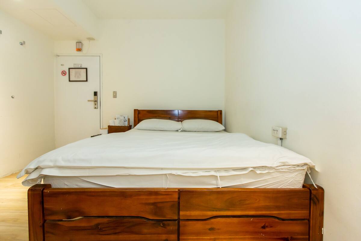 A single bed with a wooden frame and two pillows, a nightstand with a lamp, and a door with a no smoking sign.