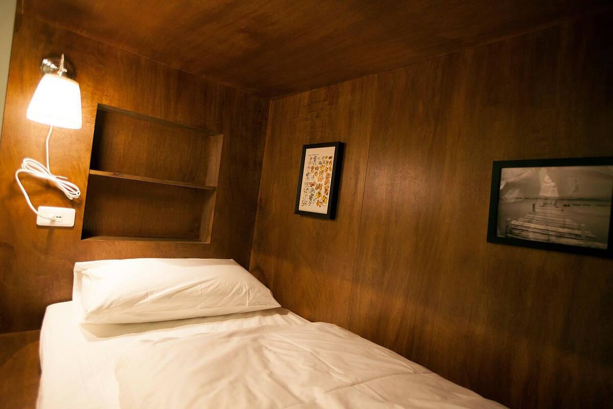 A hotel room with a wooden wall, a bed with white bedding, a bedside lamp, a shelf, and two framed pictures.