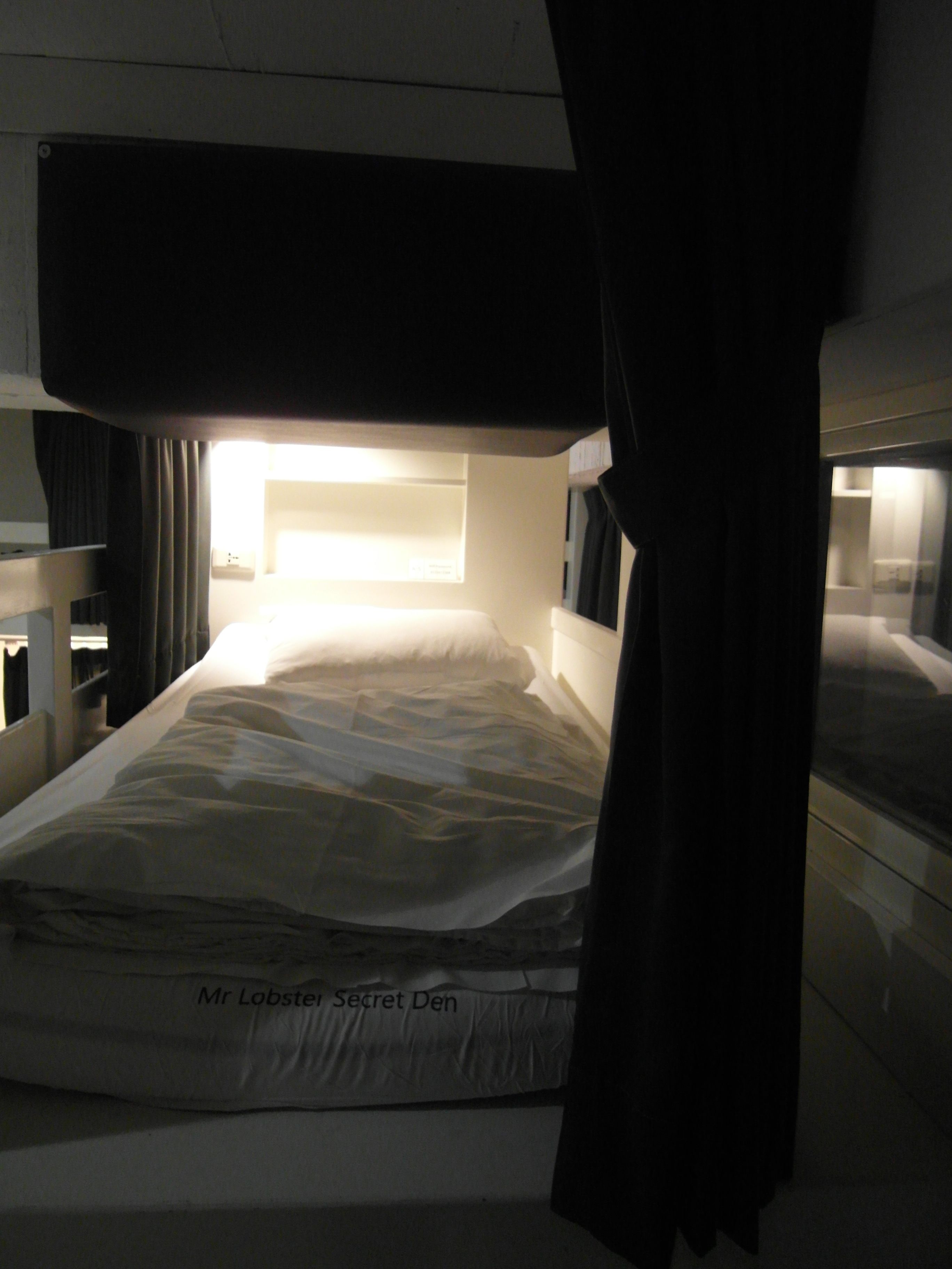 A single bed with a headboard, a bedside table, and a window with curtains.