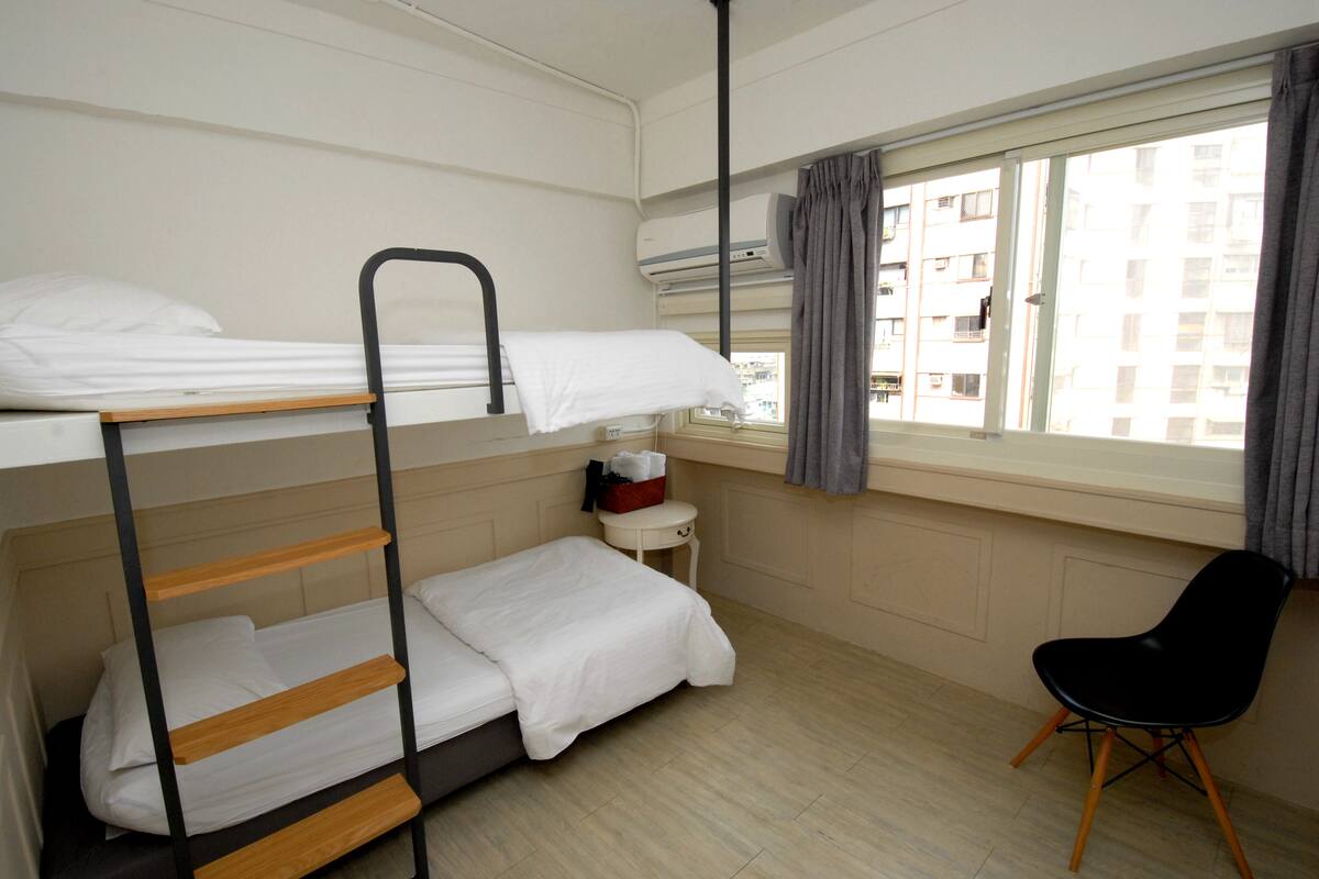 A bunk bed room with a desk and chair, a window with curtains, and a view of buildings outside.