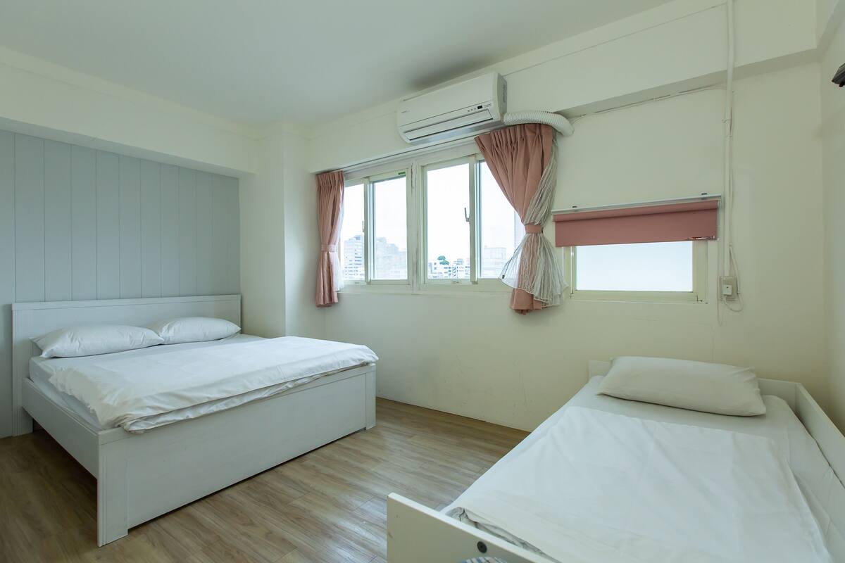 A bedroom with two beds, a window with curtains, an air conditioning unit, and a view of buildings outside.