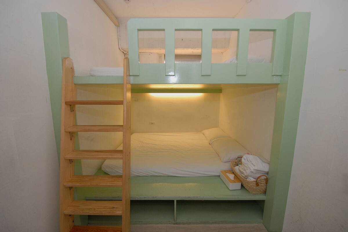 A bunk bed with a wooden ladder, a small shelf with folded towels, and a light fixture above the bed.