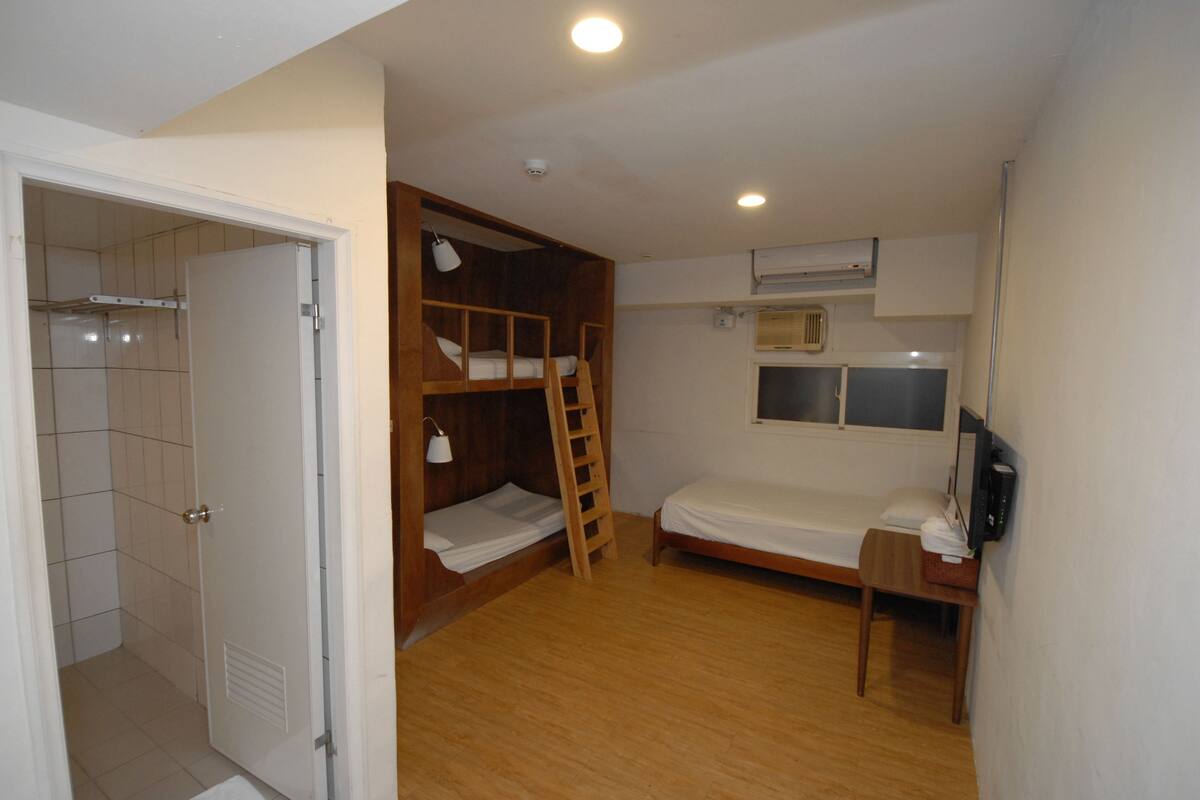 A compact room with a bunk bed, a single bed, a wooden desk, a chair, a small table, a lamp, and a TV.