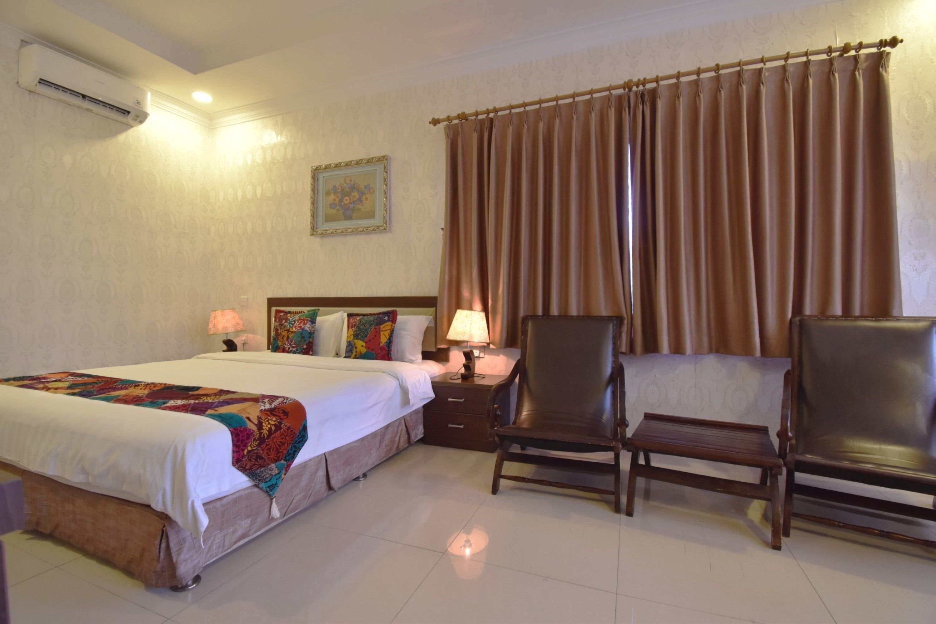 Habitación de hotel con cama, dos sillones, una mesita de noche con lámpara y ventana con cortinas.