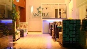 Un hall de hotel con mostrador de recepción, un letrero que dice "ANGELA" y una alfombra de bienvenida con la palabra "BIENVENIDO".