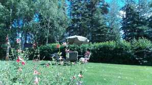 A garden with a lawn, a patio area with chairs and an umbrella, and dense trees in the background. - Fairbanks Extended Stay (Fairbanks)