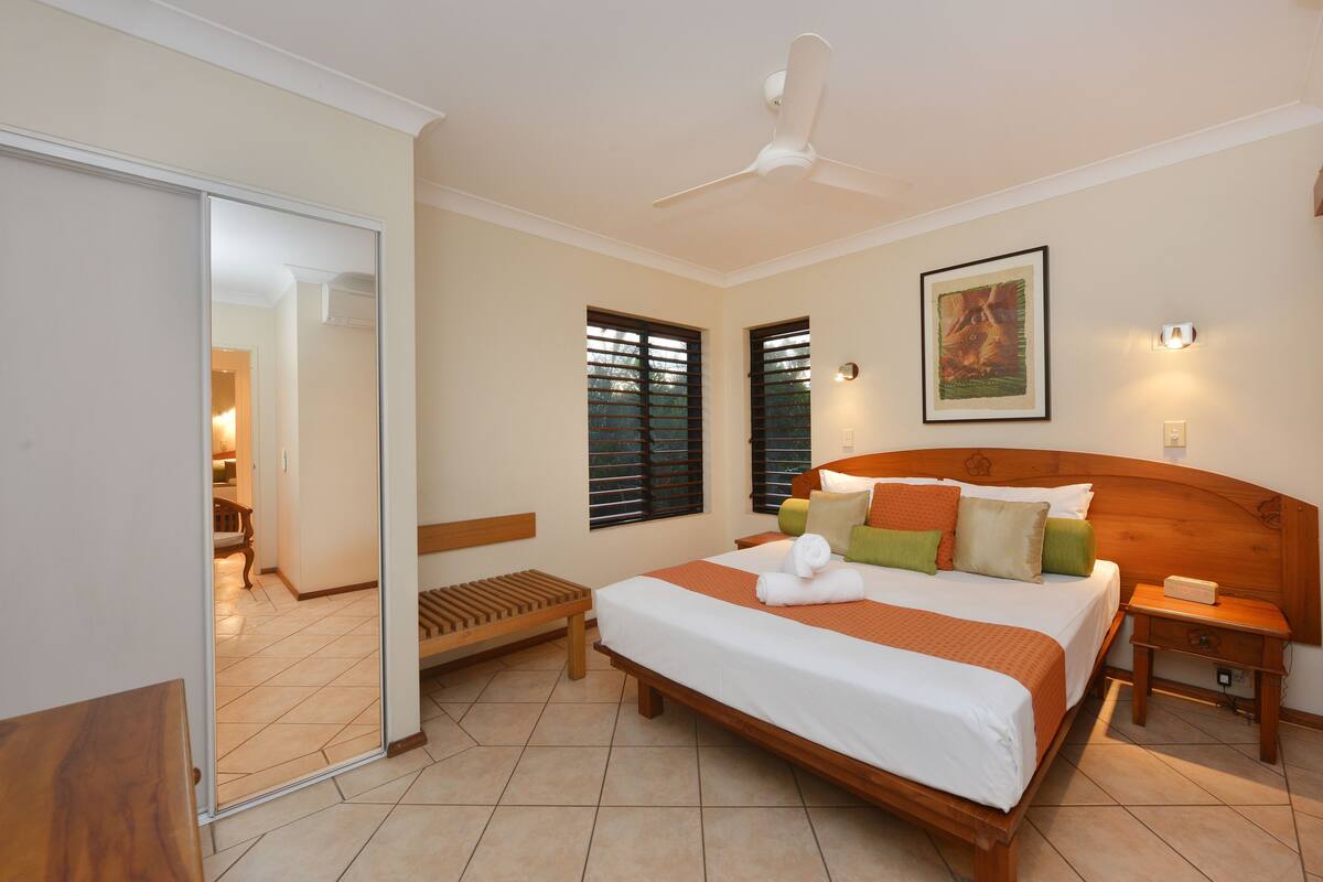 A hotel room with a large bed, a wooden headboard, a bedside table, a nightstand, a ceiling fan, and a painting on the wall.