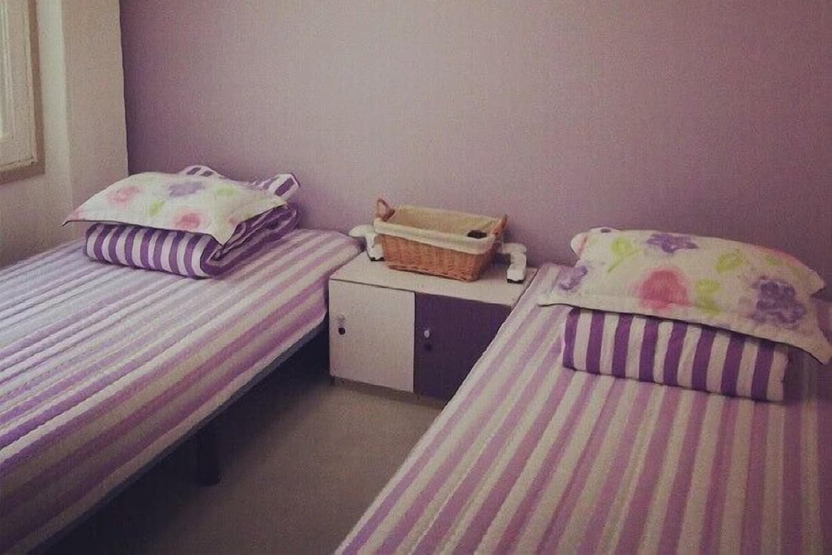 A room with two single beds, each with a striped bedspread and floral pillows. There is a small cabinet under one bed and a basket on the nightstand.