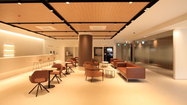 Un hall d’entrée moderne avec une colonne centrale, un espace détente avec des tables et des chaises, et un mur en verre d’un côté.
