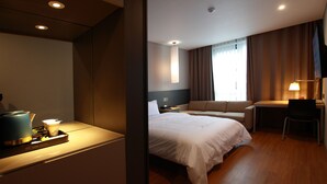 Une chambre d’hôtel comprenant un lit, un bureau, une chaise, une petite table avec une bouilloire et des tasses, ainsi qu’une fenêtre avec des rideaux.