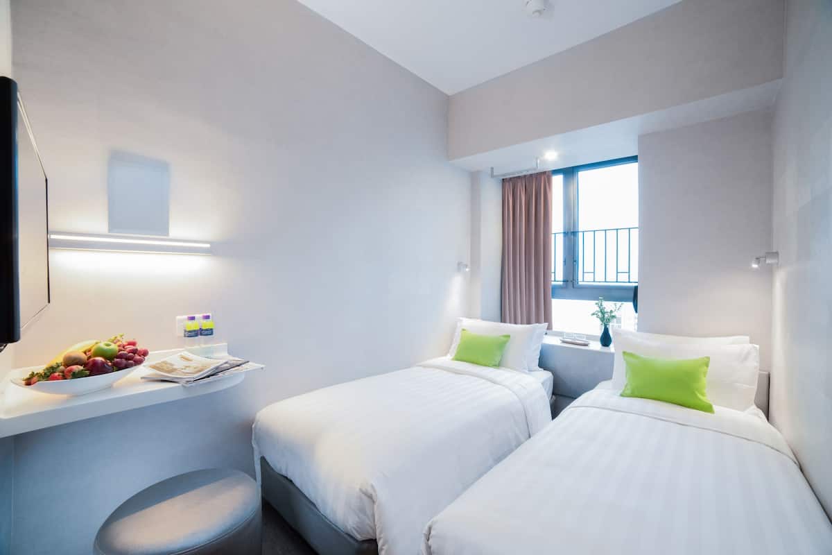 A hotel room with two beds, a small table with a fruit bowl, a mirror, and a window with curtains.