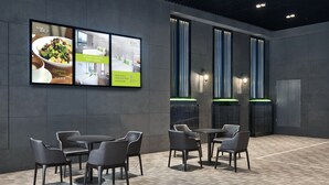 Een moderne hotellobby met een eetgedeelte met ronde tafels en stoelen, een groot digitaal scherm dat het hotel promoot, en hoge, smalle ramen met verticale jaloezieën.