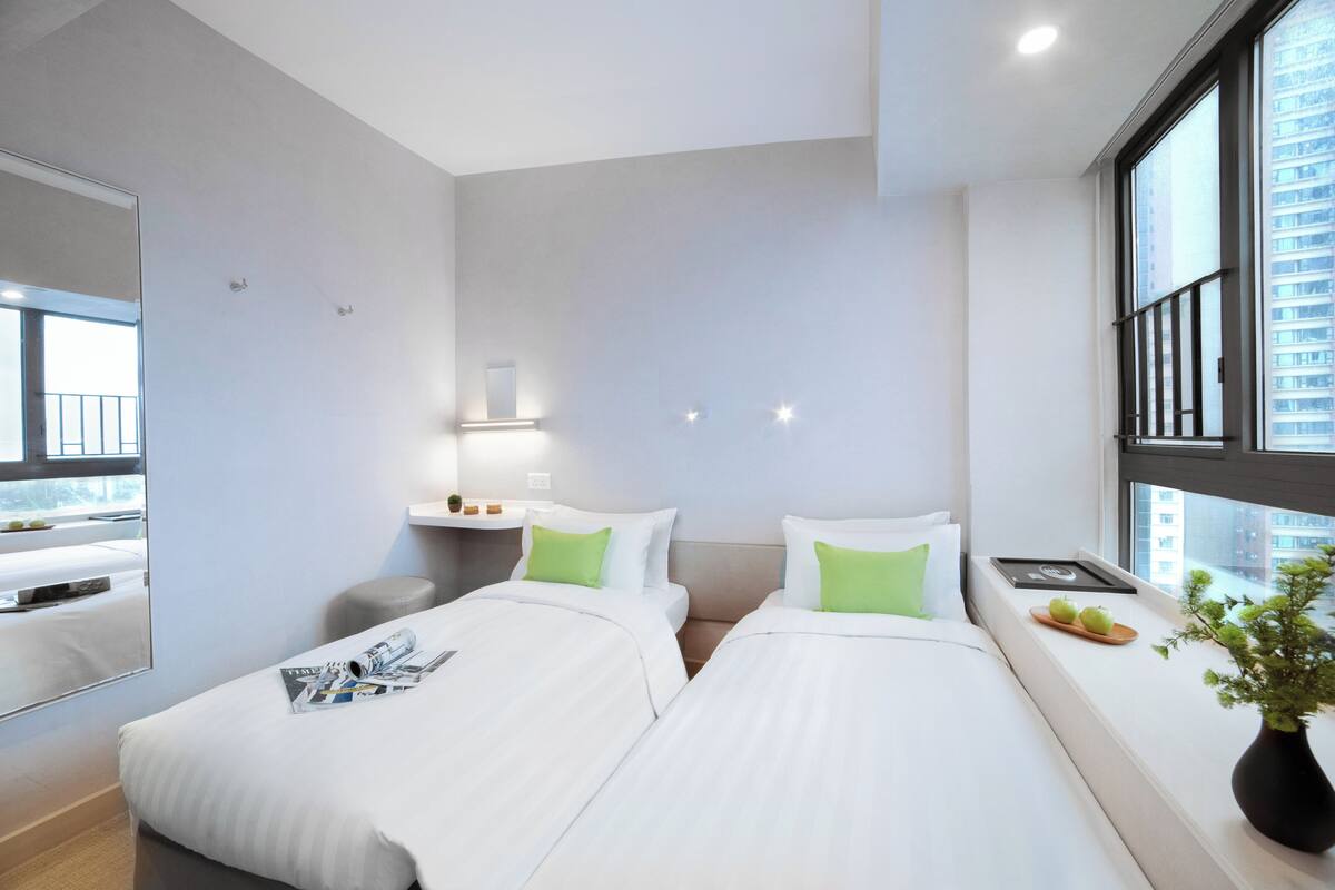 A modern hotel room with two beds, a bedside table, a nightstand, a window with a view of the city, and a potted plant on the windowsill.